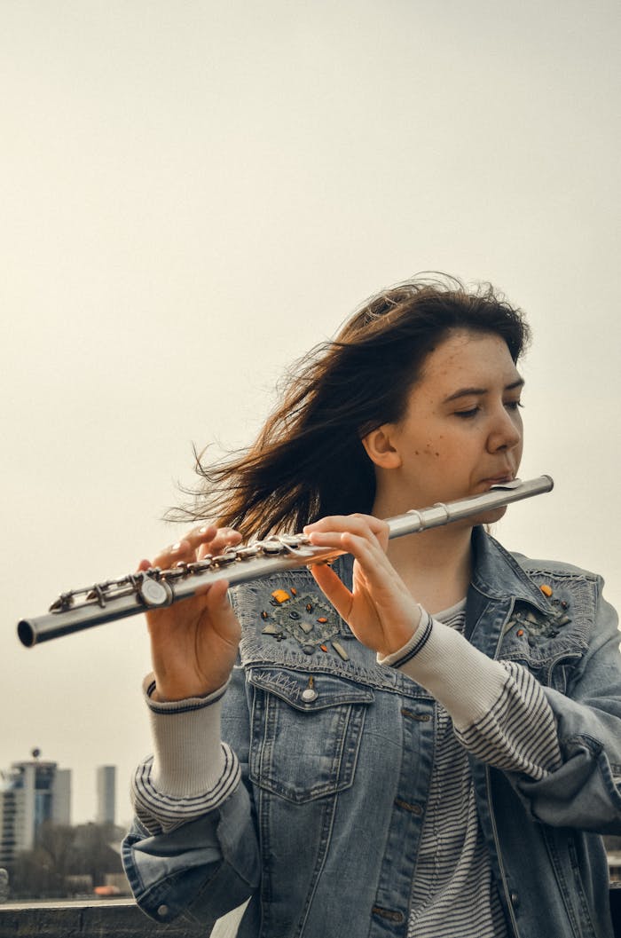 Home A young woman plays the flute outdoors, wearing a denim jacket. Captured in a serene cityscape.